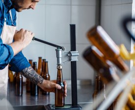 Drink production, employee works on beer manufacturing, craft plant. Young serious worker in apron closes bottles at brewery in modern plant during coronavirus quarantine, free space, cropped