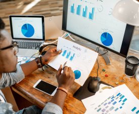 Young african man working with analitycs sitting at the beautiful working place with computer, laptop and documents. View from above